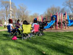 Group of park-goers sit in lawn chairs near park playground equipment.